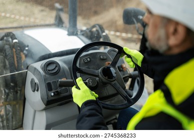 Close up of construction worker wearing protective gloves holding steering wheel inside heavy machinery cabin. Concept of control, operation and professional equipment handling.