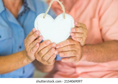 Close Up Of Couple Old Hands Holding White Wooden Heart To Represent Love And Relationship. Heart Attack Prevention Day Life Concept. Healthy Unrecognizable Mature Man And Woman