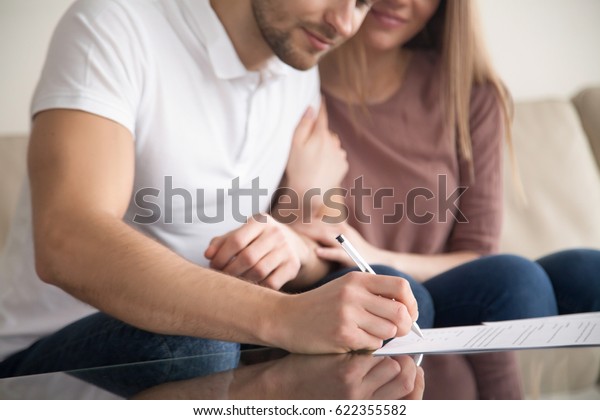 Close up of couple signing documents, young man putting\
signature on document, his wife sitting next to husband holding his\
arm, real estate purchase, first time home buyers, prenuptial\
agreement 