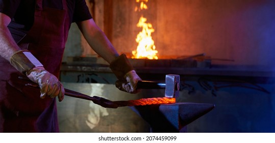 Close Up Of Craftsman In Safety Gloves Processing Heated Steel On Anvil. Strong Man Using Hammer And Forceps At Work With Metal. Burning Fire In Stove On Background.