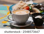 Close up of a creamy latte with delicate latte art, set on outdoor table next to a dish of grilled food. The scene captures a relaxed, sunny moment, evoking the cozy atmosphere of a casual brunch