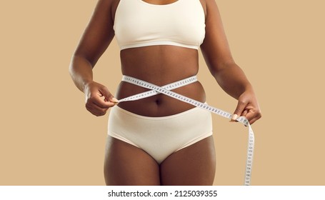 Close Up Crop Of Fat African American Woman In Underwear On Yellow Studio Background Measure Waist With Tape. Overweight Black Girl Make Measurement Before Diet. Weight Loss And Body Shape.