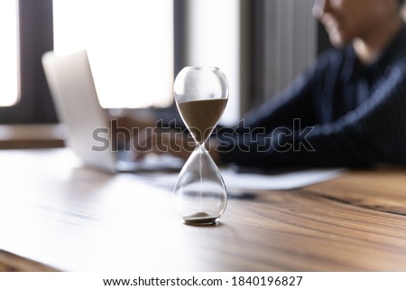 Close up crop of hourglass on wooden home office table count measure time. Female worker employee work on laptop on background, make plan or meet deadline. Time management, efficiency concept.