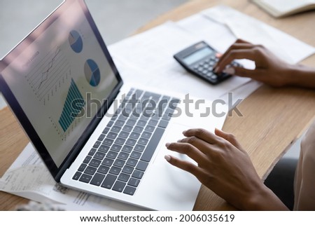 Close up cropped of African American woman working with financial project statistics, analyzing graphs on laptop screen, using calculator, calculating bills or investments, planning managing budget