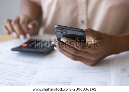 Close up cropped of African American woman using smartphone, calculating bills, browsing online banking service, checking balance, analyzing financial documents, managing planning household budget