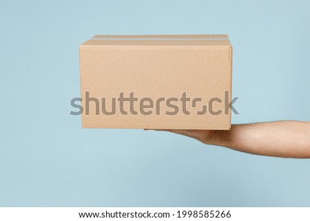Close up cropped employee courier male hold in hand brown clear blank paper empty cardboard box isolated on blue color background Packaging mock up Delivery service concept Copy space Advertising area