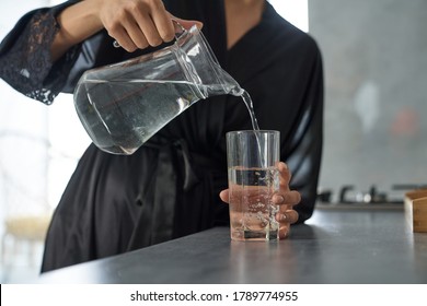 Close Up Cropped Head Of Female In Silk Dressing Gown Pouring Liquid From Pitcher Into Glass In Kitchen