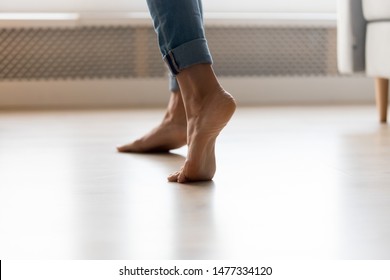Close up cropped image of barefoot girl in blue jeans standing on electric heated floor at home. Young woman stands on tiptoe, enjoy walking without room slippers on clean floor or dancing.