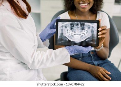 Close up cropped image of female dentist and african american woman patient at dentist's office, holding together digital tablet with patient xray panoramic jaw and teeth image. Oral care and check up