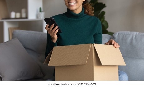 Close Up Cropped Image Laughing Happy Young Woman Sitting On Sofa With Unpacked Cardboard Parcel With Order, Leaving Positive Review In Cellphone Application, Having Good Internet Shopping Experience.