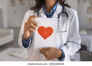 Close up cropped midsection view, unknown female doctor cardiologist wear white uniform holding postcard with drawn red heart symbol. Cardiology, regular heart check-up, healthcare, medical insurance