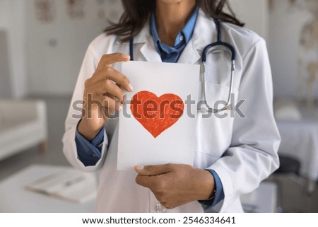Close up cropped midsection view, unknown female doctor cardiologist wear white uniform holding postcard with drawn red heart symbol. Cardiology, regular heart check-up, healthcare, medical insurance