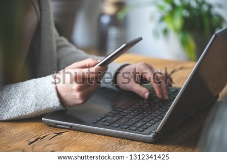 Close up and cropped photo of busy unrecognizable lady. She sitting inside loft interior co working space and using laptop, holding portable telephone equipment in her hands