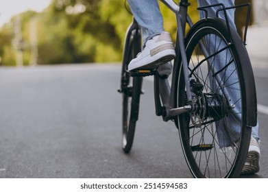 Close up cropped photo shot of young woman wearing denim pants white sneakers riding bike bicycle on road, rest relax in spring sunshine green city town park outdoors on nature. Urban leisure concept