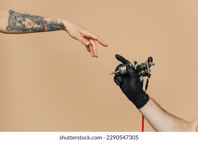 Cierre la foto recortada del artista maestro del tatuaje macho sujete con la máquina la tinta negra en el frasco, el equipo para hacer arte del tatuaje en el cuerpo, el tacto brazo del dedo hembra aislado en fondo beige claro pastel