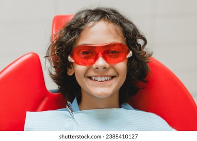 Close up cropped portrait of a child boy`s white perfect ideal toothy smile wearing protective red glasses visiting dentist orthodontist in stomatology clinic - Powered by Shutterstock