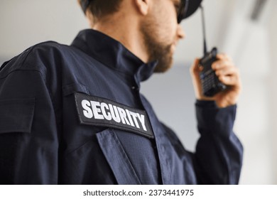 Close up cropped portrait of confident male security guard standing indoors. Bearded guardian man in blue uniform talking by the walkie talkie and looking to the side. Selective focus.