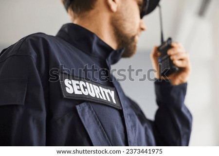 Close up cropped portrait of confident male security guard standing indoors. Bearded guardian man in blue uniform talking by the walkie talkie and looking to the side. Selective focus.