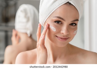 Close Up Cropped Portrait Of A Young Beautiful Woman In Bath Towel Smiling Applying Face Body Cream On Her Face For Rejuvenation Soft Moisturizing Effect. Spa Beauty Treatment Concept. Body Skin Care