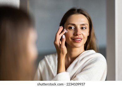 Close up cropped portrait of a young beautiful woman in bath towel smiling applying face body cream on her face for rejuvenation soft moisturizing effect. - Powered by Shutterstock