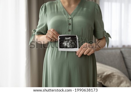 Close up cropped of pregnant woman in green dress showing ultrasound picture at camera, standing at home, young future mom demonstrating scan, waiting for first baby, pregnancy and motherhood concept