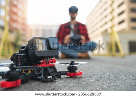 Close up cropped shot shot of generic desing fpv drone after landing on a street road, with male pilot on the background. Unmanned aviation, operating fpv multicopter experience concept. 