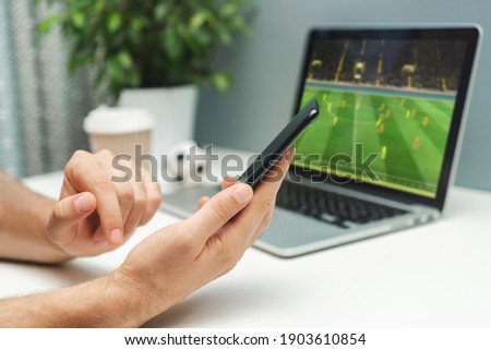 Close up cropped shot of male hand holding smartphone. Man watching soccer play live broadcast online on his laptop and making bets on his favourite team using mobile application.