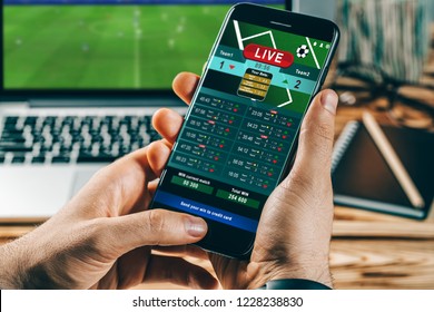 Close up cropped shot of male hands making bets using gambling mobile application on his phone. Man watching football match online broadcast on his laptop waiting for winning results.  - Shutterstock ID 1228238830