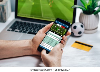 Close up cropped shot of male hands making bets using gambling mobile application on his phone. Man watching football match online broadcast on his laptop waiting for winning results. - Shutterstock ID 1569629827