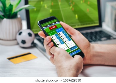 Close up cropped shot of male hands making bets using gambling mobile application on his phone. Man watching football match online broadcast on his laptop waiting for winning results. - Shutterstock ID 1569629830