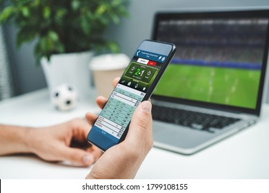 Close Up Cropped Shot Of Male Hands Betting Online Using Gambling Mobile Application On His Phone. Man Watching Football Match Online Broadcast On His Laptop Waiting For Winning Results.