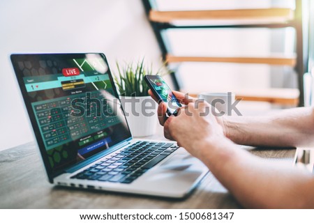 Close up cropped shot of male hands making bets using gambling mobile application on his phone. Man watching football match online broadcast on his laptop waiting for winning results.