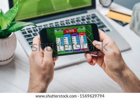 Close up cropped shot of male hands making bets using gambling mobile application on his phone. Man watching football match online broadcast on his laptop waiting for winning results.

