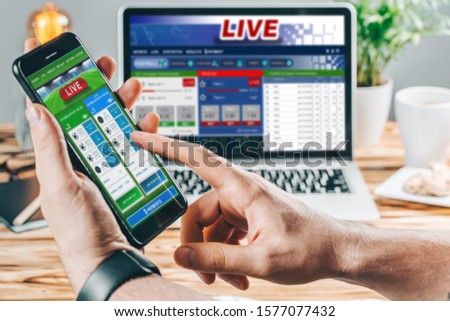 Close up cropped shot of male hands betting online using gambling mobile application on his phone. Man watching football match online broadcast on his laptop waiting for winning results.