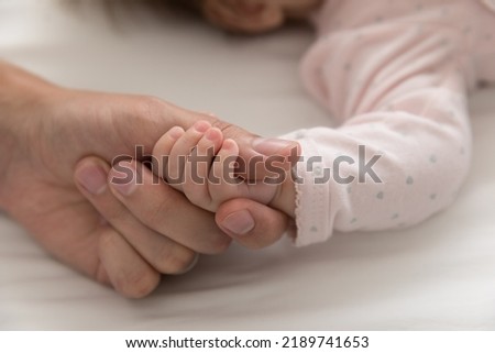 Close up cropped shot of tiny arm of cute newborn, baby girl holds daddy thumb during carefree healthy sleep in bed. Family bond and ties, parent unconditional love, cherish, happy paternity concept
