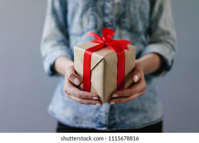 Close up cropped shot of a woman dressed in denim shirt  holding gift box wrapped in craft paper and decorated with red satin ribbon in her hands. Plain grey background.