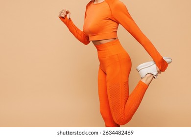 Close up cropped side view young fitness trainer instructor sporty woman wear orange clothes spend time in gym raise up leg stretch isolated on plain beige background. Workout sport fit abs concept