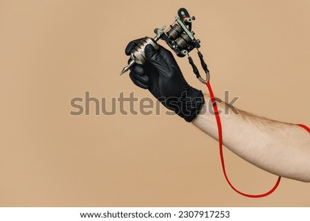 Close up cropped tattooer master artist male hold in hand machine black ink in jar, equipment for making tattoo art on body isolated on pastel plain beige background. Copy space advertising mock up