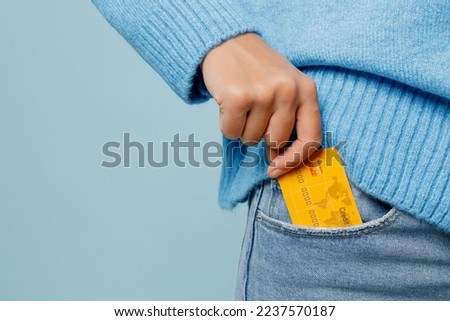 Close up cropped woman wearing knitted sweater hold in hand put into pocket mock up of credit bank card isolated on plain pastel light blue cyan background studio portrait. People lifestyle concept