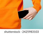 Close up cropped young man he wears orange casual clothes hold in hand put into pocket mobile cell phone with blank screen isolated on plain blue cyan color wall background studio. Lifestyle concept