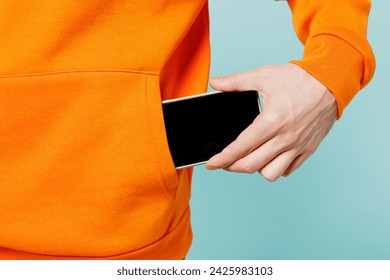 Close up cropped young man he wears orange casual clothes hold in hand put into pocket mobile cell phone with blank screen isolated on plain blue cyan color wall background studio. Lifestyle concept