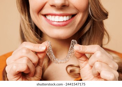 Close up cropped young woman wear orange shirt casual clothes hold in hand invisible transparent aligners, invisalign bracer smile isolated on plain pastel light beige background. Lifestyle concept - Powered by Shutterstock