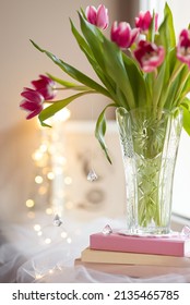 Close Up Of A Crystal Vase With A Bouquet Of Pink Tulips On A Stack Of Books On A Window Sill With Blurred Fairy Lights In The Background