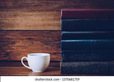 Close up of a cup of coffee with stack of old book on wood table over wooden wall background
