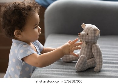 Close up curious cute toddler African American girl touching stuffed toy bear profile portrait, standing near cozy couch at home, pretty little child playing in playroom, childcare concept - Powered by Shutterstock