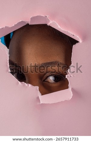 Close up of a curious dark skinned model peering through a ripped pink backdrop, creating a captivating and mysterious portrait