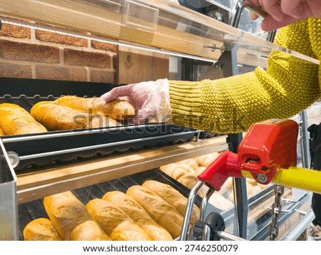 Close up of customer using plastic glove to take fresh baguette from bakery display in supermarket. Grocery shopping concept with freshly baked bread in self service store aisle