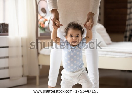 Close up cute African American toddler girl taking first steps, looking at camera, holding caring young mother hands, loving mum supporting adorable little daughter, teaching to walk, childcare