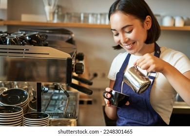 Close up of cute asian barista girl making cappuccino, doing latte art in cup with steamed milk, standing in coffee shop behind counter. - Powered by Shutterstock