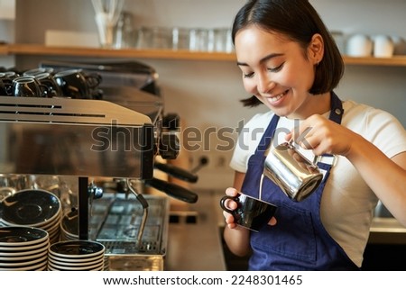 Close up of cute asian barista girl making cappuccino, doing latte art in cup with steamed milk, standing in coffee shop behind counter.